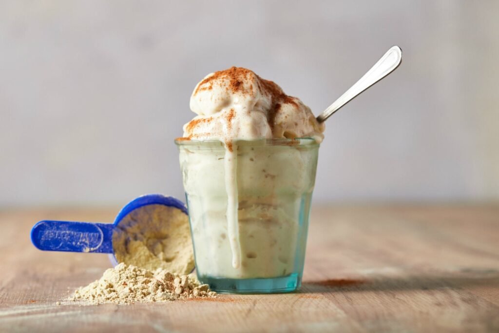 A creamy serving of homemade protein ice cream in a glass, topped with a sprinkle of cinnamon, with a scoop of protein powder next to it. A healthy, high-protein dessert option made using casein protein powder for a rich texture.