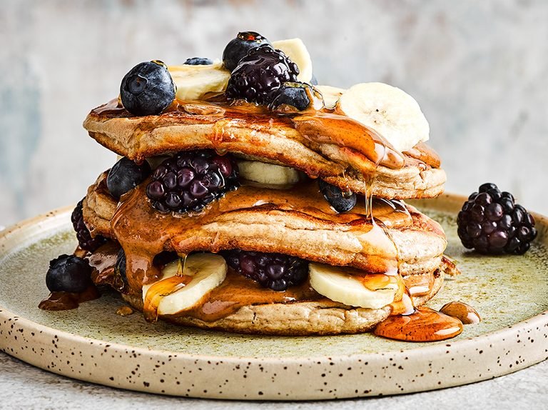 Stack of fluffy protein pancakes topped with fresh blackberries, blueberries, banana slices, and drizzled with syrup, served on a rustic plate.